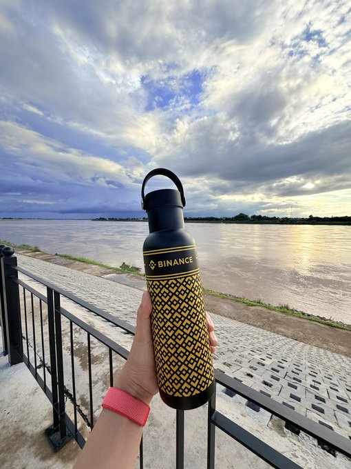 A hand wearing a pink watch band holds up a black and yellow Binance-branded water bottle against a scenic riverside view with cloudy skies at sunset.