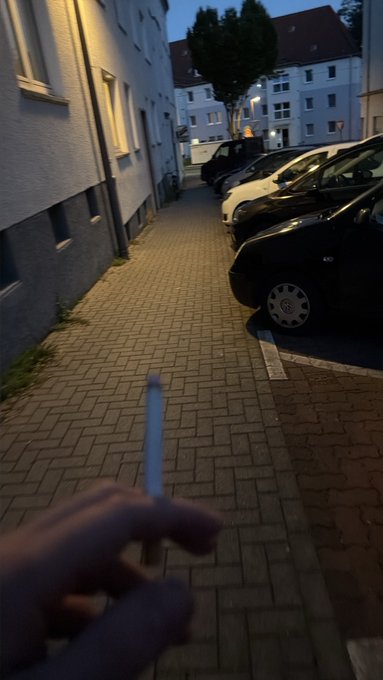 A first-person perspective showing a hand holding a cigarette, walking down a brick-paved street with parked cars at dusk.