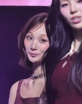 Two women with long dark hair are shown in a dimly lit setting with a purple tint, wearing reddish-burgundy outfits.