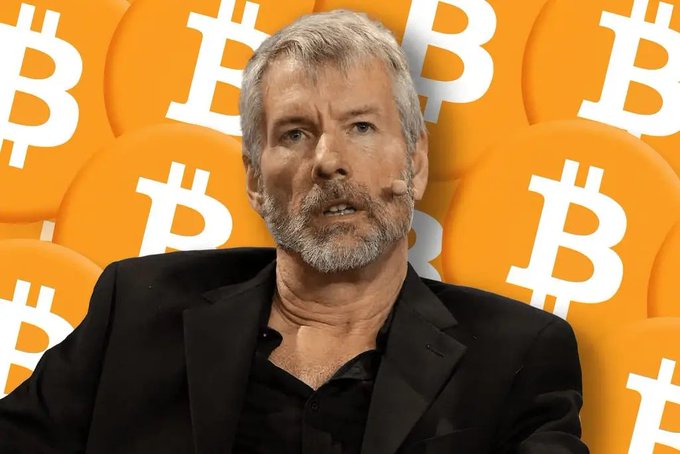 A man with grey hair and a beard speaks in front of a backdrop featuring orange Bitcoin logos.