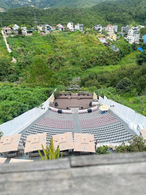 An outdoor amphitheater stage set up on a hillside, with rows of empty chairs, beige umbrellas, and residential buildings visible in the green, mountainous background.