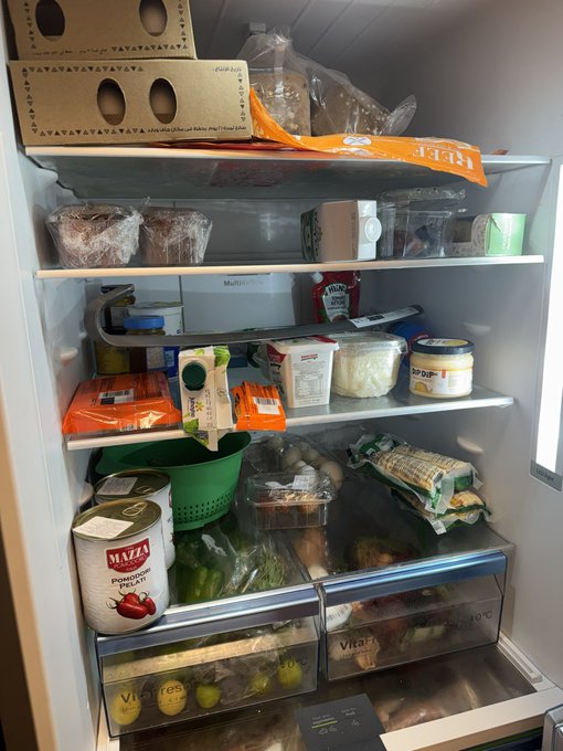 This photo shows a crowded refrigerator filled with various groceries, including Mazza tomato cans, bread, and various containers of food. The image illustrates the social media prompt by providing a candid look into the owner's eating habits and lifestyle through their food choices.