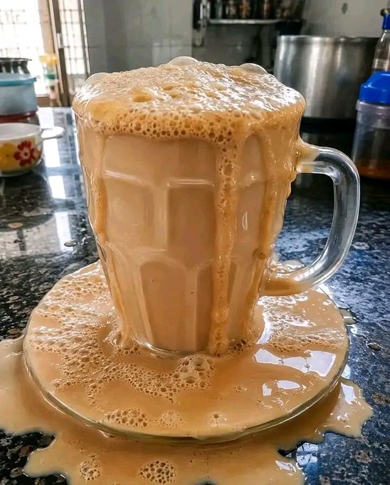 A glass mug overflowing with frothy milky tea sits on a saucer, with the liquid spilling over the rim and pooling across a dark granite countertop.