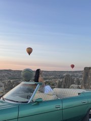 A woman in a turquoise vintage convertible car takes in the breathtaking view of hot air balloons floating over the rocky landscape of Cappadocia, Turkey, at sunrise—a unique travel moment that contrasts with the post's reflection on staying committed to the crypto space through its "ups and downs" alongside Binance.