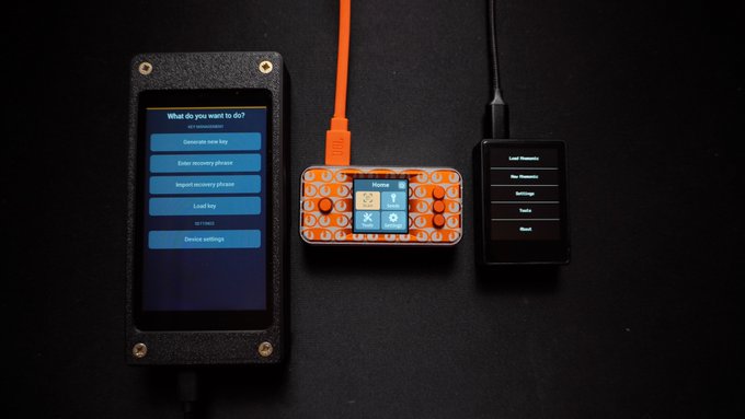 Three electronic devices laid out on a dark surface: a black device with a touchscreen showing key management options, an orange handheld gaming-style device, and a smaller screen displaying a menu.