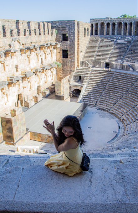 A woman in a flowing yellow dress sits on ancient stone steps at a Roman amphitheater, smiling and posing with her arms raised, with the curved stone seating tiers visible behind her.