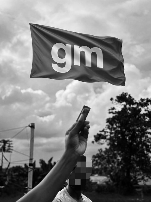 A black-and-white image of a person waving a flag emblazoned with "gm" against a cloudy sky, capturing the upbeat "good morning" spirit of the post.
