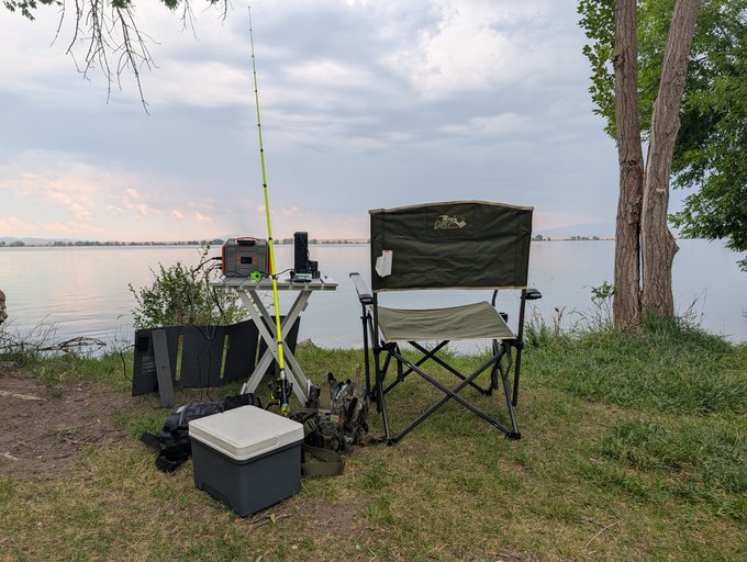A fishing setup on a grassy shore by a lake at dusk, featuring a green folding chair, a small table with tackle, a fishing rod with a bright yellow-green pole, and a white cooler.