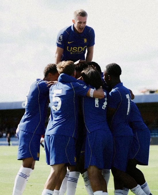 A group of soccer players in blue Chelsea uniforms celebrating a goal, with players jumping on each other in a huddle on a sunny field.