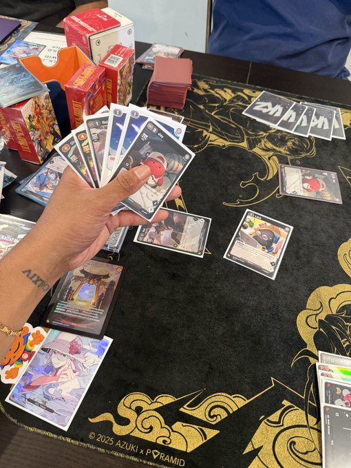 A person holding a fan of trading cards at a gaming event, with booster packs, a deck box, and a playmat featuring gold designs visible on the table.