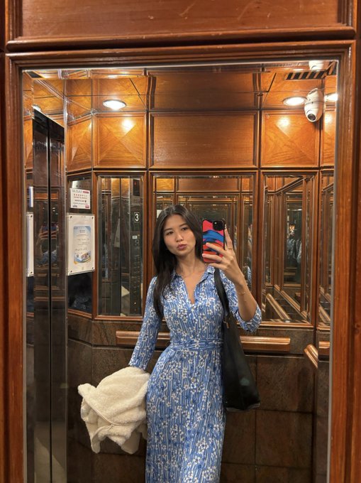 A woman takes a mirror selfie in an ornate wooden-paneled elevator while wearing a blue floral midi dress, holding a white fuzzy jacket and a black shoulder bag. The post's "gm" (good morning) message and "aths" (all-time highs) reference align with her confident, smiling morning look.