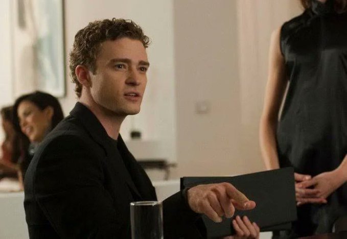 A young man with curly hair in a black blazer points while holding a menu at a restaurant, with other patrons visible in the background.