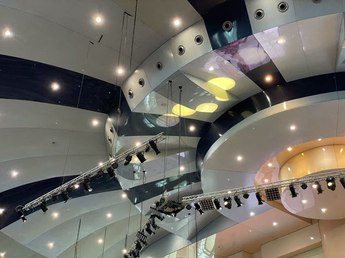 An interior view looking up at a modern building's ceiling with curved white panels, recessed lighting, and hanging lighting trusses rigged with stage lights.