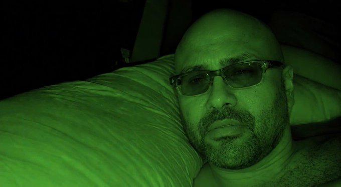 A bald man with glasses and a beard lies on a pillow, captured in a selfie with a green night-vision tint.