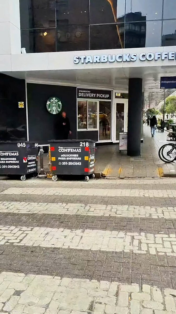 The image shows a Starbucks coffee shop with a person standing outside, possibly waiting for someone or just passing by. The scene also includes a black suitcase near the entrance of the store, which could be related to an ongoing delivery or transportation issue. Additionally, there is a bicycle parked nearby and a car in front of the building.