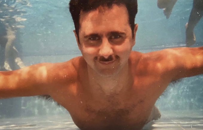 A shirtless man with dark hair and a mustache is underwater in a swimming pool, arms outstretched toward the camera in a carefree pose, capturing the "log off" essence of the caption. The image, likely from the 1980s based on its film quality, accompanies the post's sentiment of stepping away from crypto markets to embrace a moment of leisure.