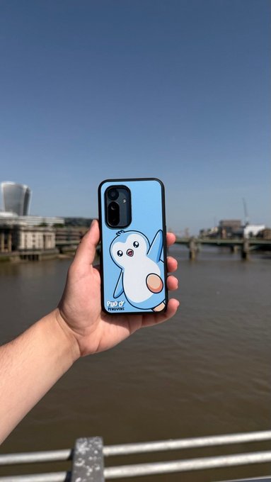 A hand holds a phone with a blue phone case featuring a cartoon white bear-like creature, set against a backdrop of a river and city skyline.
