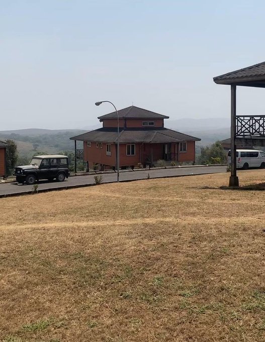 A scenic outdoor view shows a red-roofed house on a hill with a vintage Land Rover parked in the driveway and a grassy lawn in the foreground.