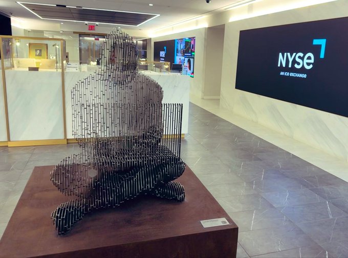 A metal sculpture of a seated figure sits in front of an illuminated "NYSE An ICE Exchange" sign at the New York Stock Exchange, with a small plaque at its base identifying it as the Bitcoin statue.