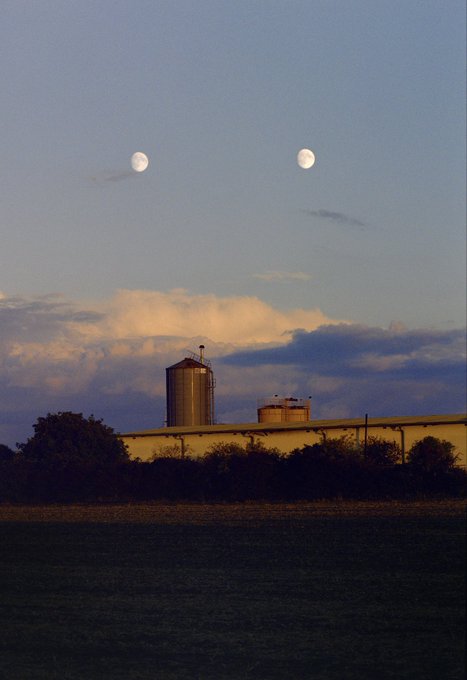A dusk landscape showing two moons in the sky above industrial silos and a long building, with trees silhouetted in the foreground.