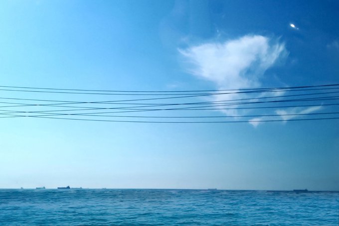 A bright blue sky with wispy white clouds stretches over a calm ocean, with power lines crossing the frame and ships visible on the distant horizon.