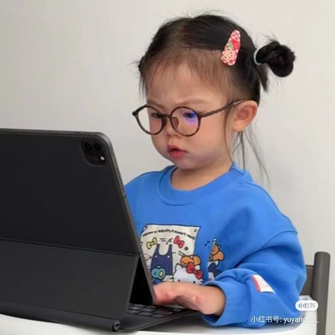 A young child wearing round glasses and a blue Hello Kitty sweatshirt intently uses a tablet with an attached keyboard, appearing focused on screen work. The image humorously illustrates the "back to the grind" message, depicting an adorable toddler resembling a studious worker diligently engaging with their device.