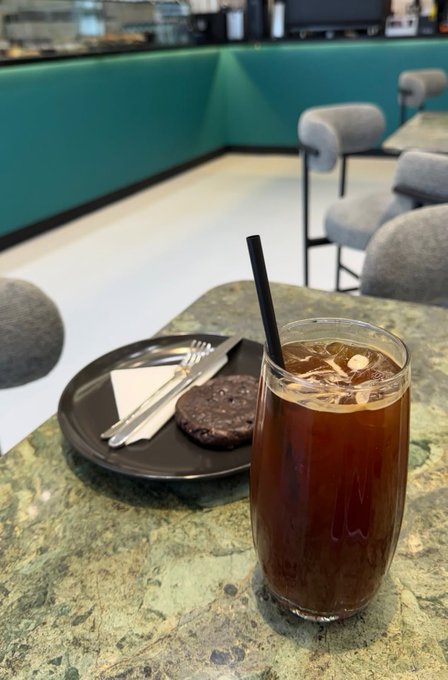 A glass of iced coffee with a black straw sits on a green marble table, accompanied by a black plate with a cookie, fork, and napkin in a cafe setting.