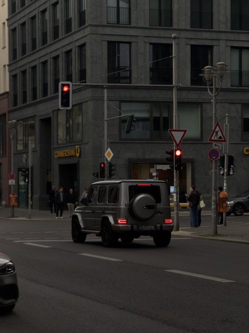 A silver Mercedes-Benz G-Class SUV drives on a city street past a Commerzbank building, with red traffic lights and pedestrians visible at the intersection.