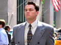 Leonardo DiCaprio in a gray suit and patterned tie, standing on a city street with an American flag visible in the background.