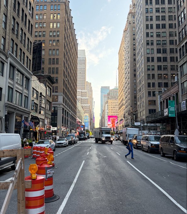A daytime city street view in what appears to be New York, featuring tall buildings on both sides, construction barriers with orange barrels in the foreground, and various vehicles including trucks and vans on the road.