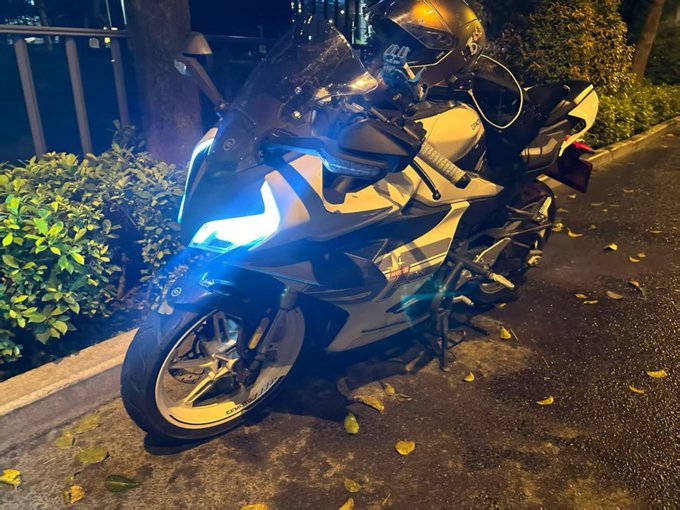 A sleek sportbike with a helmet resting on it is parked on a leaf-strewn street at night, its bright LED headlight glowing. The rider appears to have ditched the bicycle plan in favor of taking out their motorcycle instead.