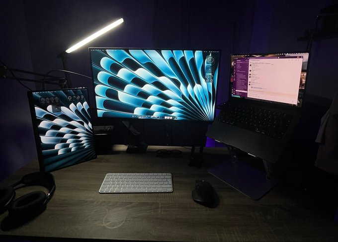 A dimly lit desk setup featuring a vertical monitor, a main curved display, and a laptop on a stand, all illuminated by a bright LED light bar and showing matching blue abstract wallpapers.
