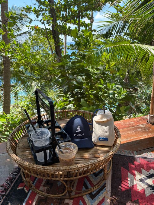 A cozy outdoor scene featuring a rattan table set with iced coffees and a "PHAROS" branded cap alongside a hydration flask, set against a lush tropical backdrop with ocean views—capturing the essence of "RealFi, IRL" by showcasing a tangible, real-world moment of financial independence through leisure and travel.