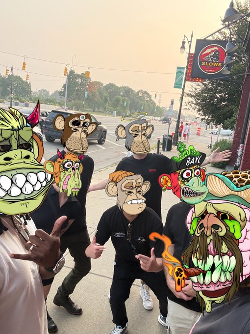 A group of people on a city street wearing black shirts pose with cartoonish monster masks and graphics, with a 'SLOW'S' BBQ sign visible on a building in the background.