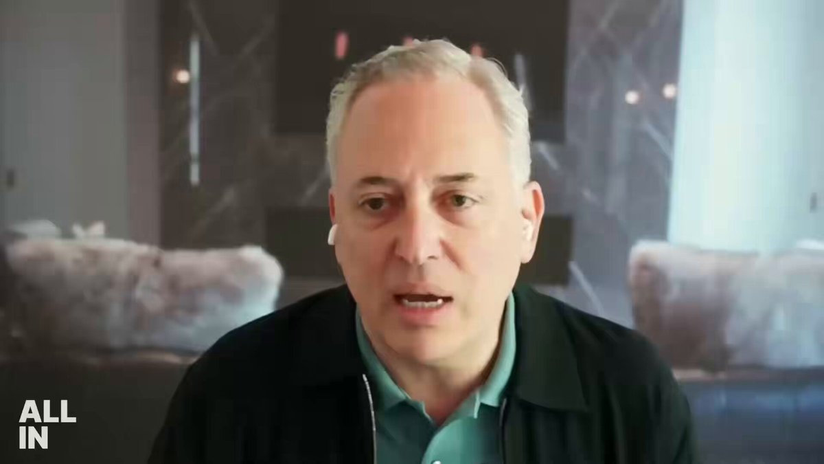The image features a man wearing a black jacket, sitting in front of a fireplace with a gray couch behind him. He appears to be talking or giving a speech while looking directly at the camera. The caption below the image reads "All In".