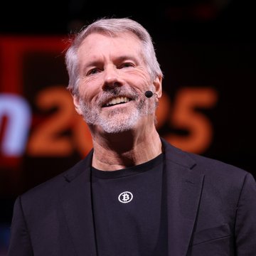 The image features a man wearing a black suit jacket and speaking into a microphone while standing in front of a large screen displaying text related to Bitcoin transactions. The man appears to be giving a speech or presentation, possibly discussing the value of Bitcoin or its potential future. The background shows an orange sign with white text that reads "2x2".