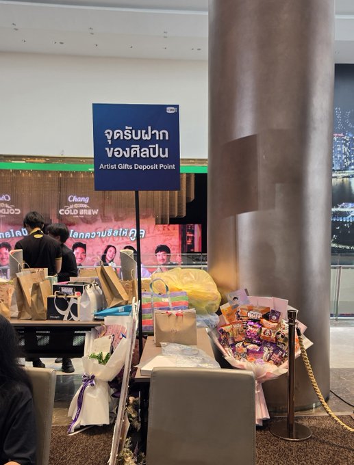 A photo of an 'Artist Gifts Deposit Point' (จุดรับฝากของศิลปิน) at an event, displaying tables full of snacks, gifts, and bouquets of flowers.