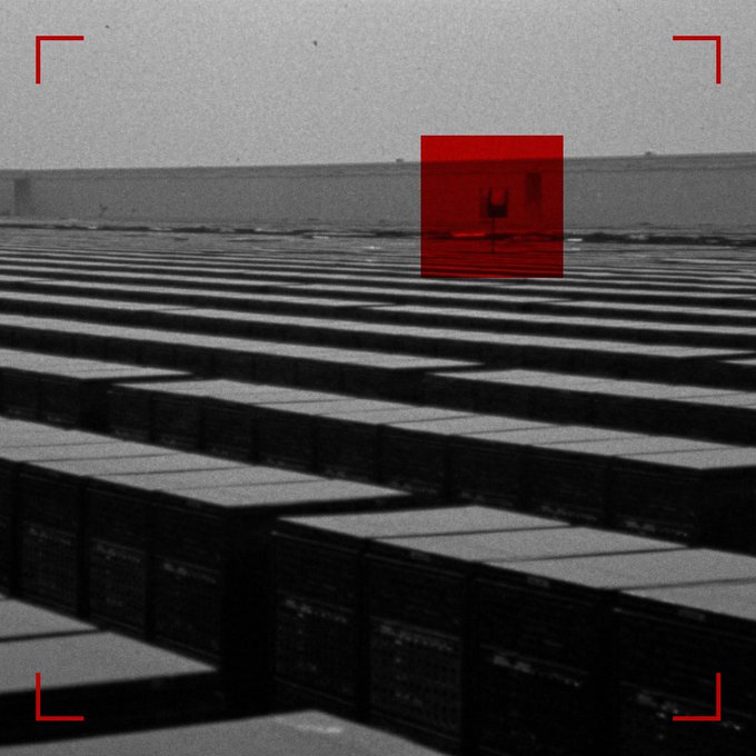 A stylized grayscale image with red camera viewfinder brackets and a red highlight box, showing rows of what appears to be servers or industrial containers with a small figure in the distance.