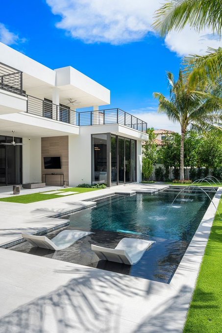 A photograph of a modern white two-story house featuring a swimming pool with built-in lounge chairs, palm trees, and a covered outdoor patio area.