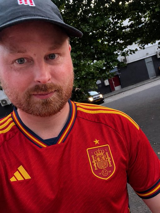 A bearded man wearing a cap and a red Spain national football team Adidas jersey takes a selfie on a street with trees and a car visible in the background.