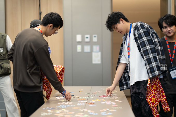 A group of young men gathered around a table in a room, focused on sorting or arranging a large collection of small stickers.