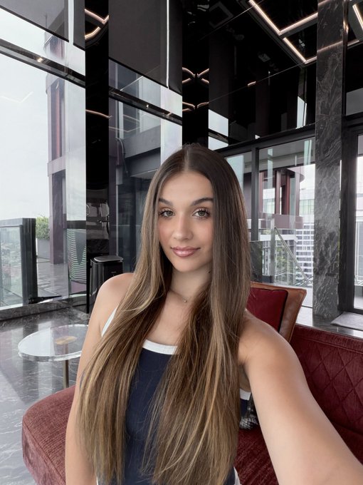 A young woman with long brown hair takes a selfie in a modern, upscale interior with large windows and contemporary decor.