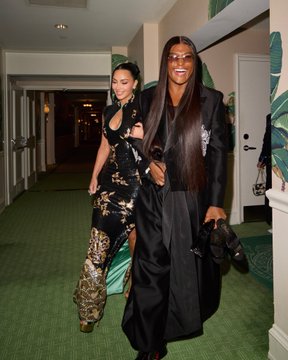 A photo of two women walking down a hallway with green carpet; one wears a snakeskin-patterned outfit and the other wears an oversized black blazer and sunglasses.