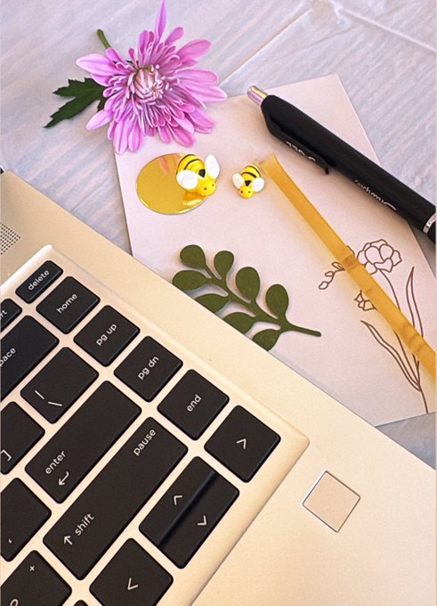 A flat lay photograph showing a laptop keyboard, a pink chrysanthemum flower, a notebook with bee stickers, and pens arranged on a light wooden surface.