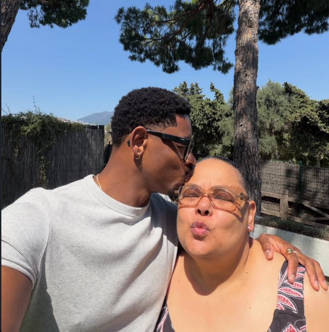 Selfie of a young man in sunglasses kissing an older woman on the cheek while outdoors near trees and a wooden fence.