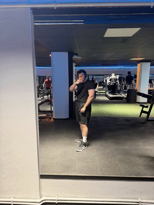A mirror selfie of a man in a black t-shirt and shorts holding a phone in a gym with treadmills and blue-lit pillars in the background.