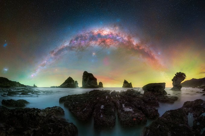 The image shows a beautiful night sky with a large, colorful galaxy in the center of the frame. The galaxy appears to be floating above some rocks near the ocean, creating an interesting contrast between the natural elements and the celestial spectacle. The photo is taken from a low angle, which emphasizes the grandeur of the galaxy against the backdrop of the dark sky.