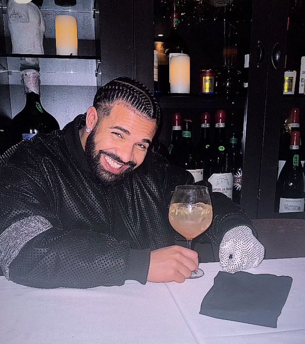 A man with cornrow braids and a beard smiles broadly while holding a cocktail glass in front of a shelf of wine bottles at a restaurant, the scene visually celebrating the massive $12.5M slot win referenced in the post.