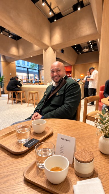 A man smiles at a wooden table in a stylish café, with two pour-over coffee setups and a small card reading "WatchHouse" visible, fitting the post about his visit to WatchHouse NYC on 5th Ave. The image captures the relaxed moment of enjoying coffee at his favorite spot.