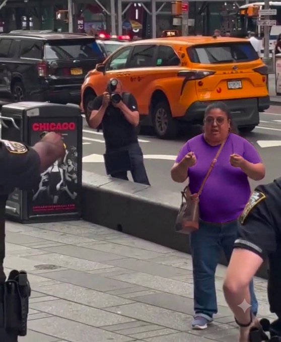 A street scene in Chicago showing a woman in a purple shirt gesturing animatedly while a person with a camera films her, with an orange taxi and a 'CHICAGO' newspaper box visible.